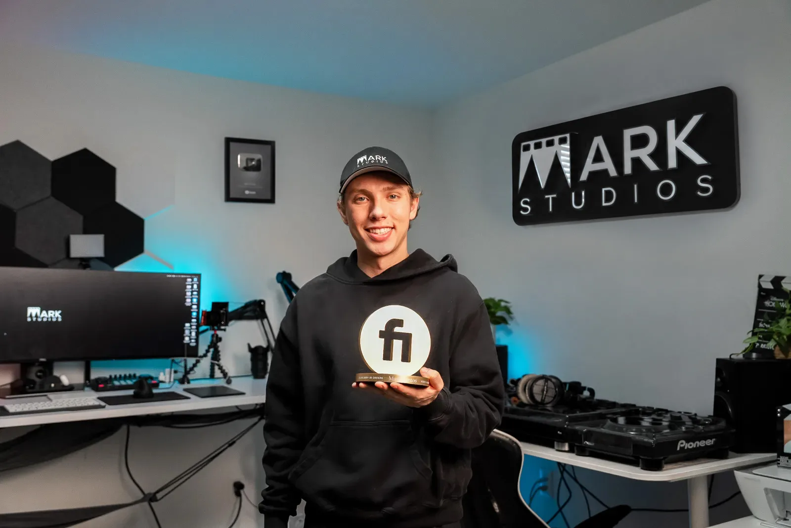 Mark Santos in the Mark Studios office holding Fiverr's gold award, with the Mark Studios sign and his YouTube Silver Play Button on the wall behind him